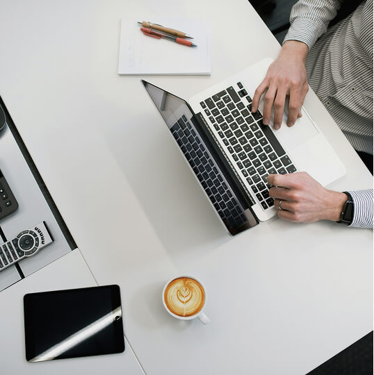 ノートパソコンで作業する人物の手元と、デスクに置かれたコーヒーとタブレット