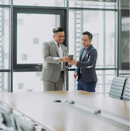 ガラス張りの会議室で、スマートフォンを見ながら話している2人の人物